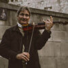 smiling standing man playing violin by gray stone wall